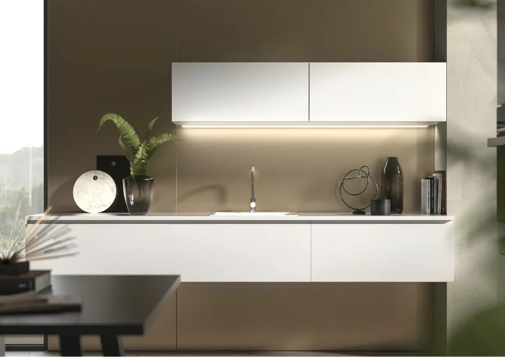 L-shaped kitchen with sleek white cabinets, soft underlighting and minimalist styling against a warm beige wall