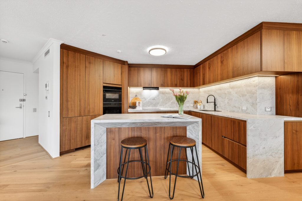 Luxury Häcker kitchen featuring fluted walnut timber cabinetry and premium beige marble bench tops, splashback, and waterfall kitchen island.